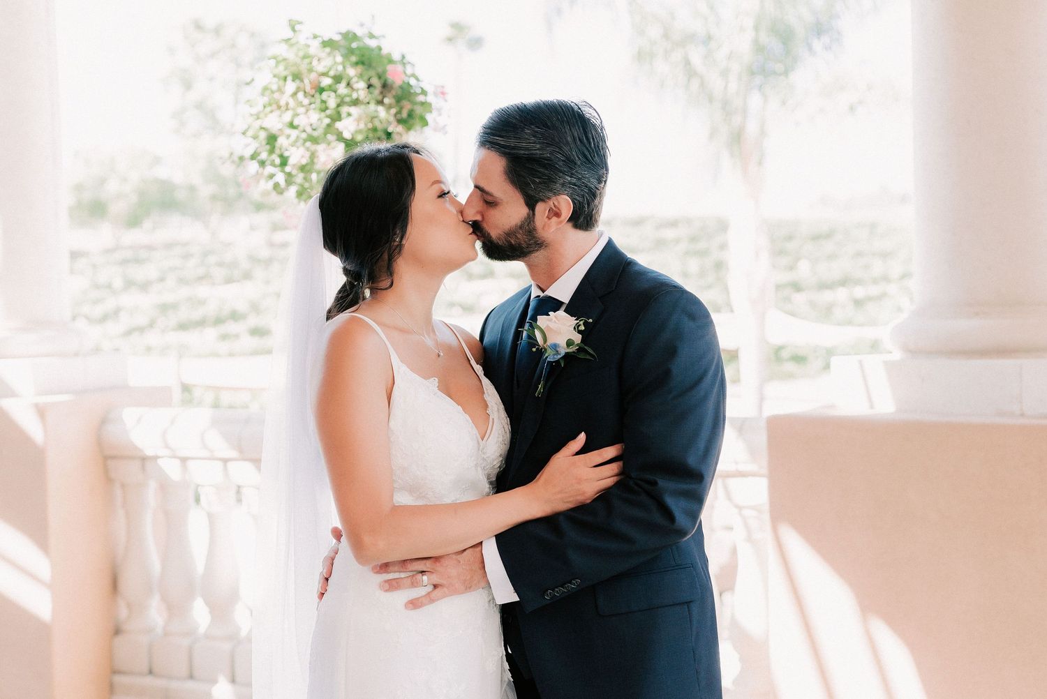 A bride and groom share a romantic kiss at an elegant outdoor venue, dressed in a white gown and navy suit.