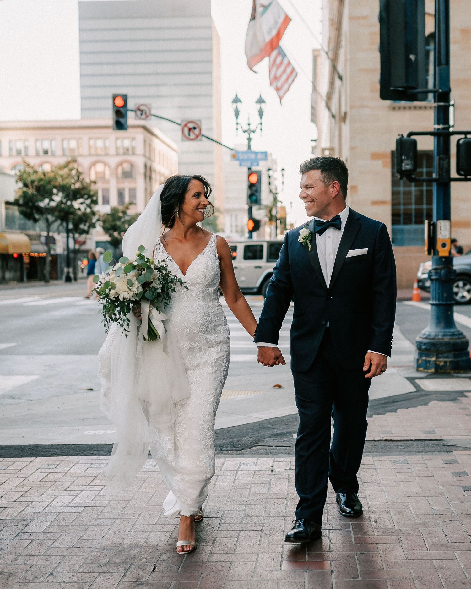 Bride and groom holding hands walking on a city street, bride in white gown with bouquet, groom in black tuxedo.