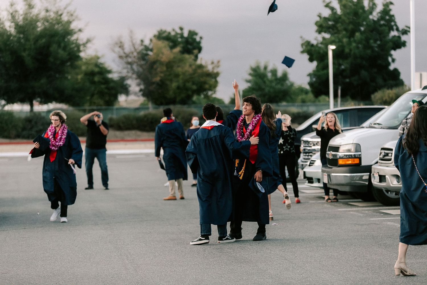 Graduates in black robes toss caps in a parking lot, celebrating their graduation ceremony outdoors.