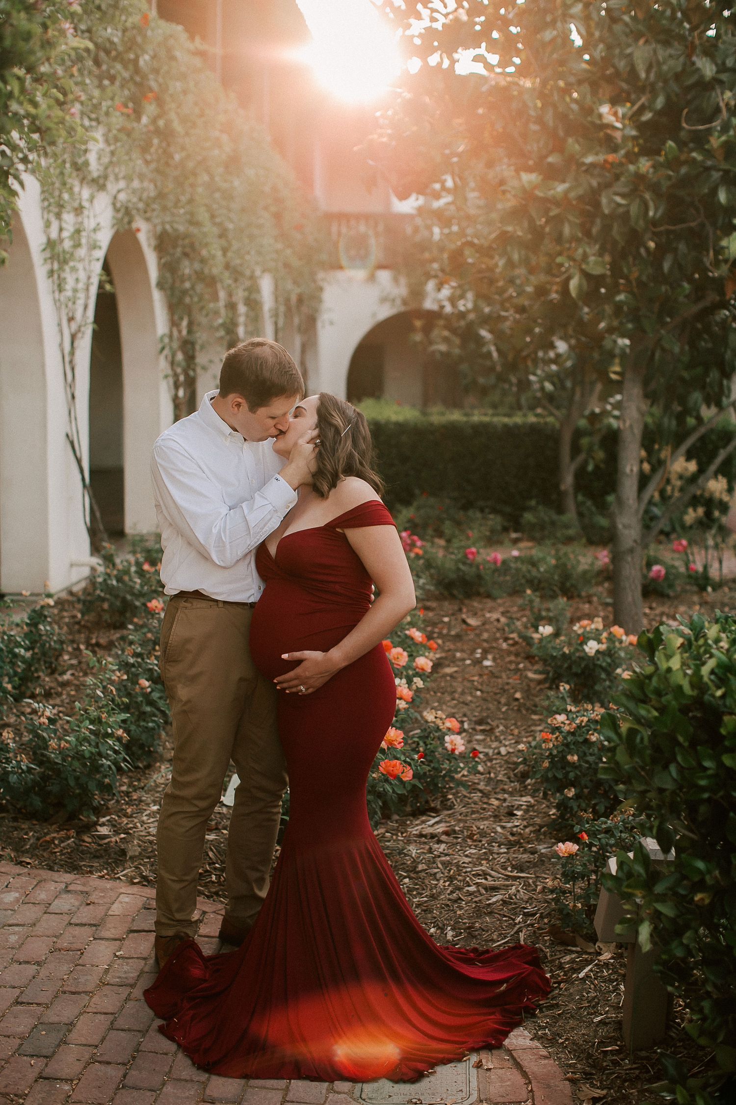 Couple sharing a kiss in a garden, pregnant woman in a flowing red off-shoulder gown, golden sunset light behind them.