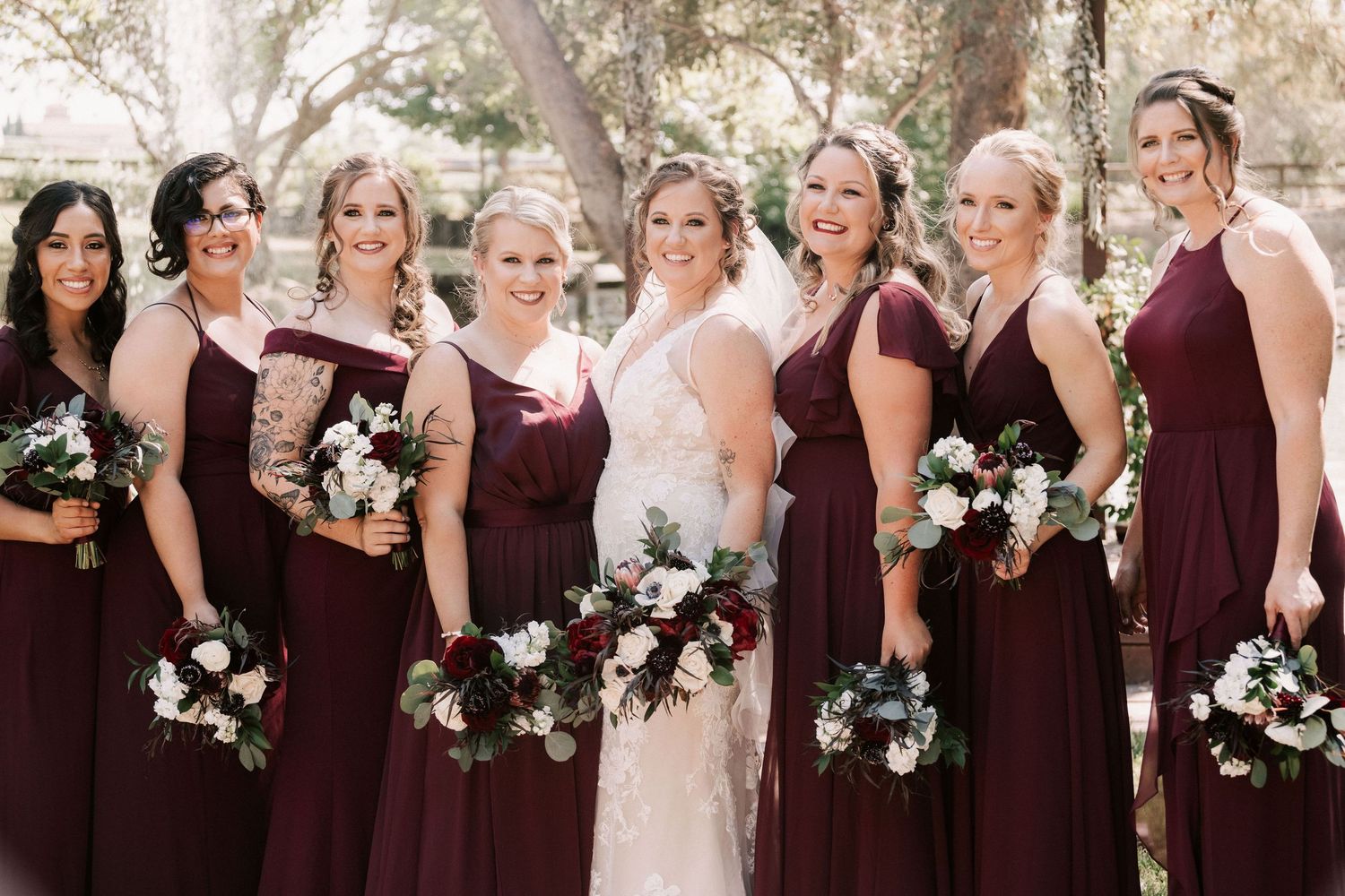 Bride in lace gown poses with six bridesmaids in burgundy dresses holding floral bouquets outdoors.