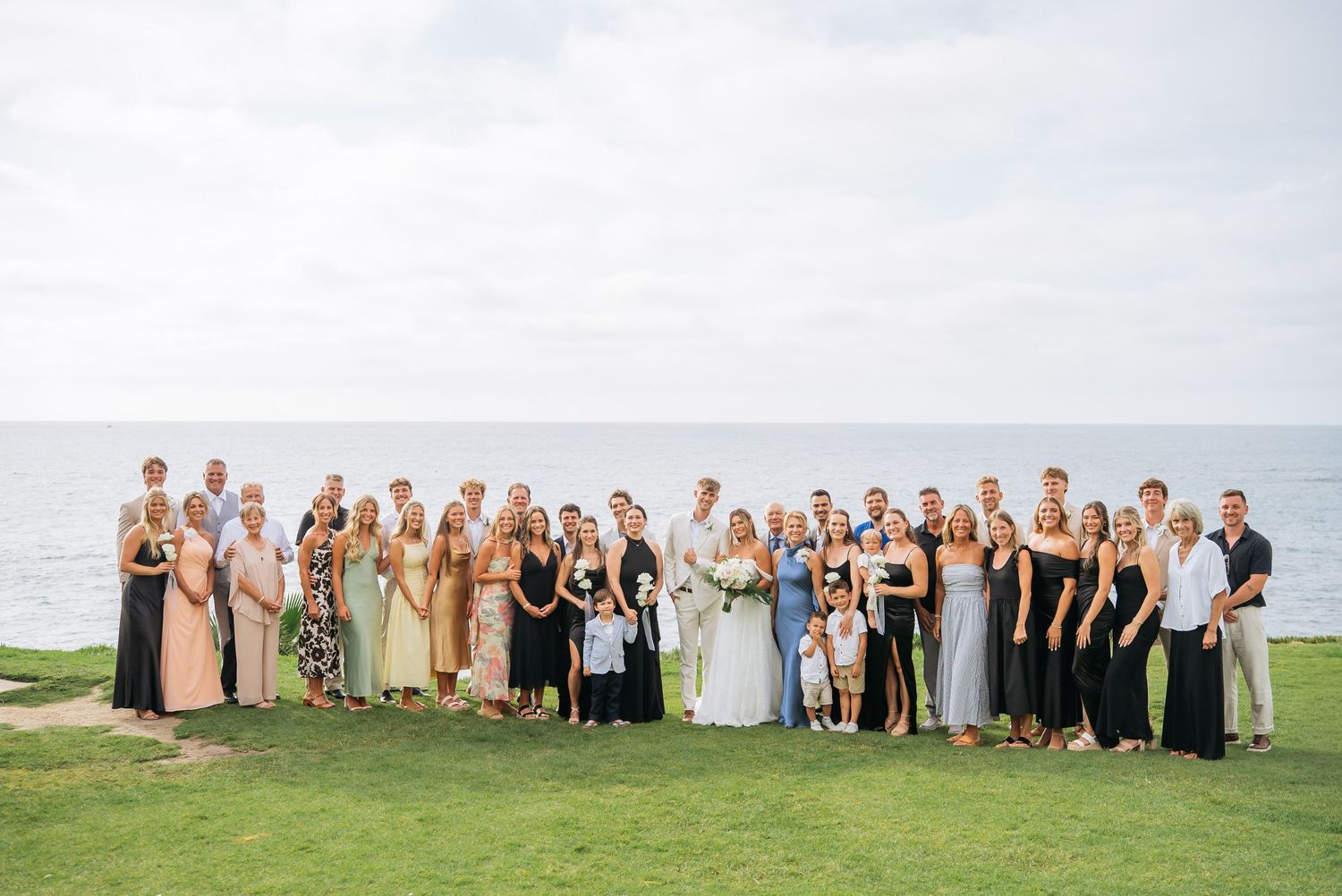 Large wedding party group photo outdoors on green lawn with ocean backdrop on a cloudy day.