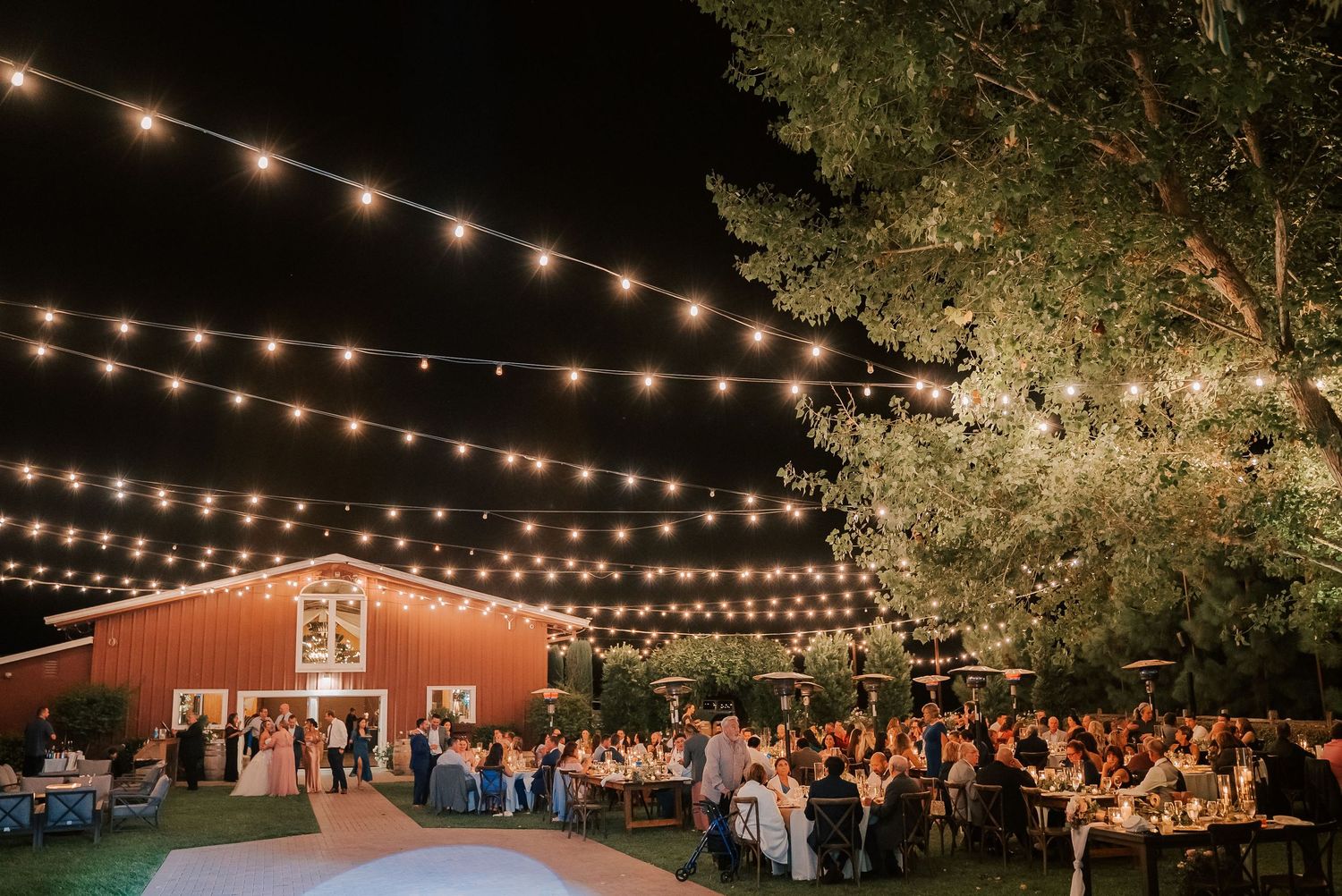 Outdoor barn wedding reception at night with string lights illuminating guests dining under trees beside a red barn.