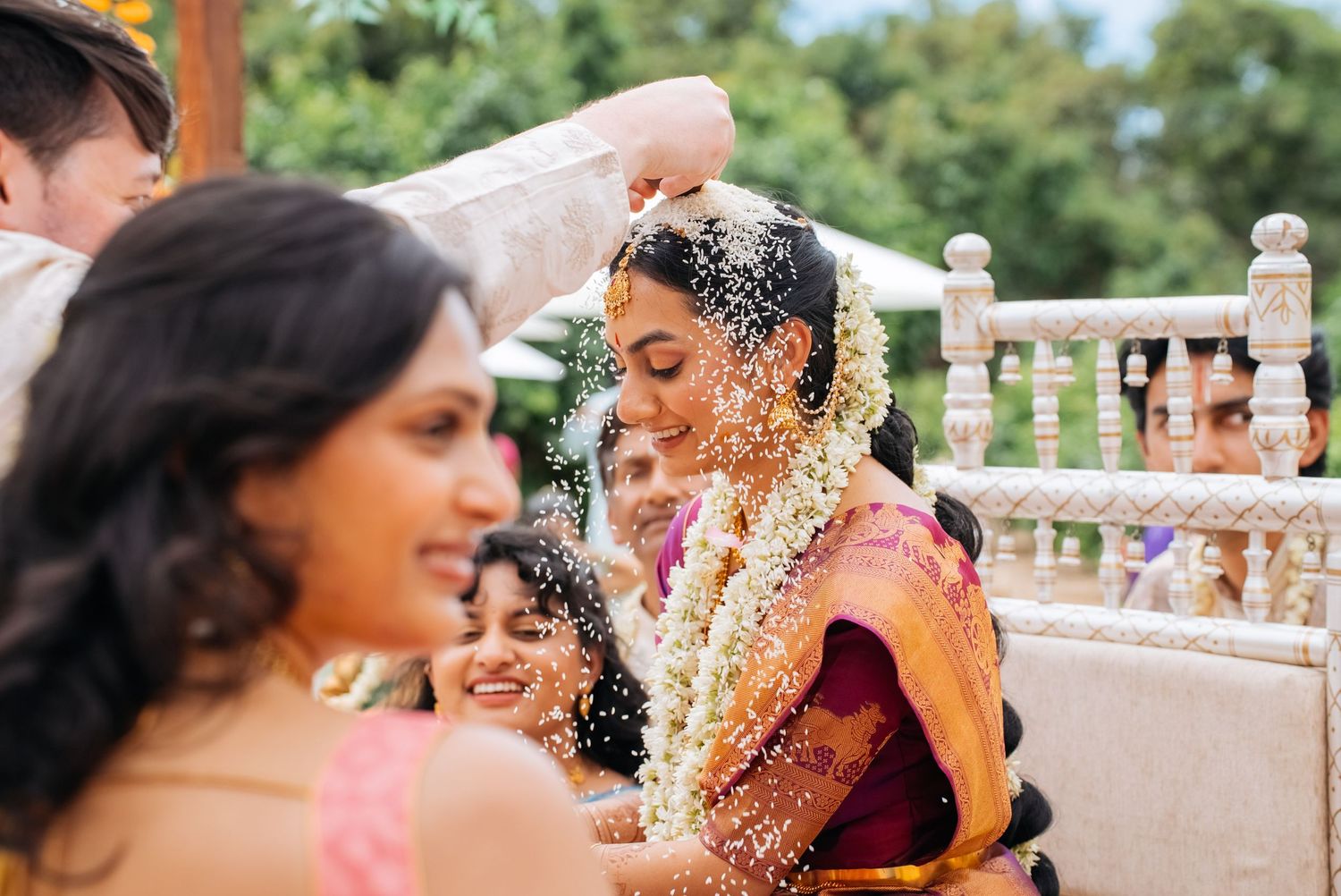 A bride in a red saree adorned with jasmine garlands receives rice blessings during a traditional South Indian wedding ceremony.