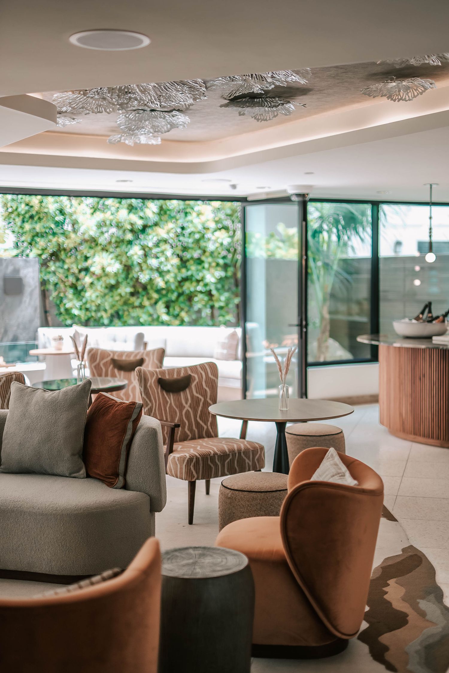 Elegant hotel lounge with terracotta chairs, gray sofa, decorative ceiling art, and floor-to-ceiling windows overlooking lush greenery.