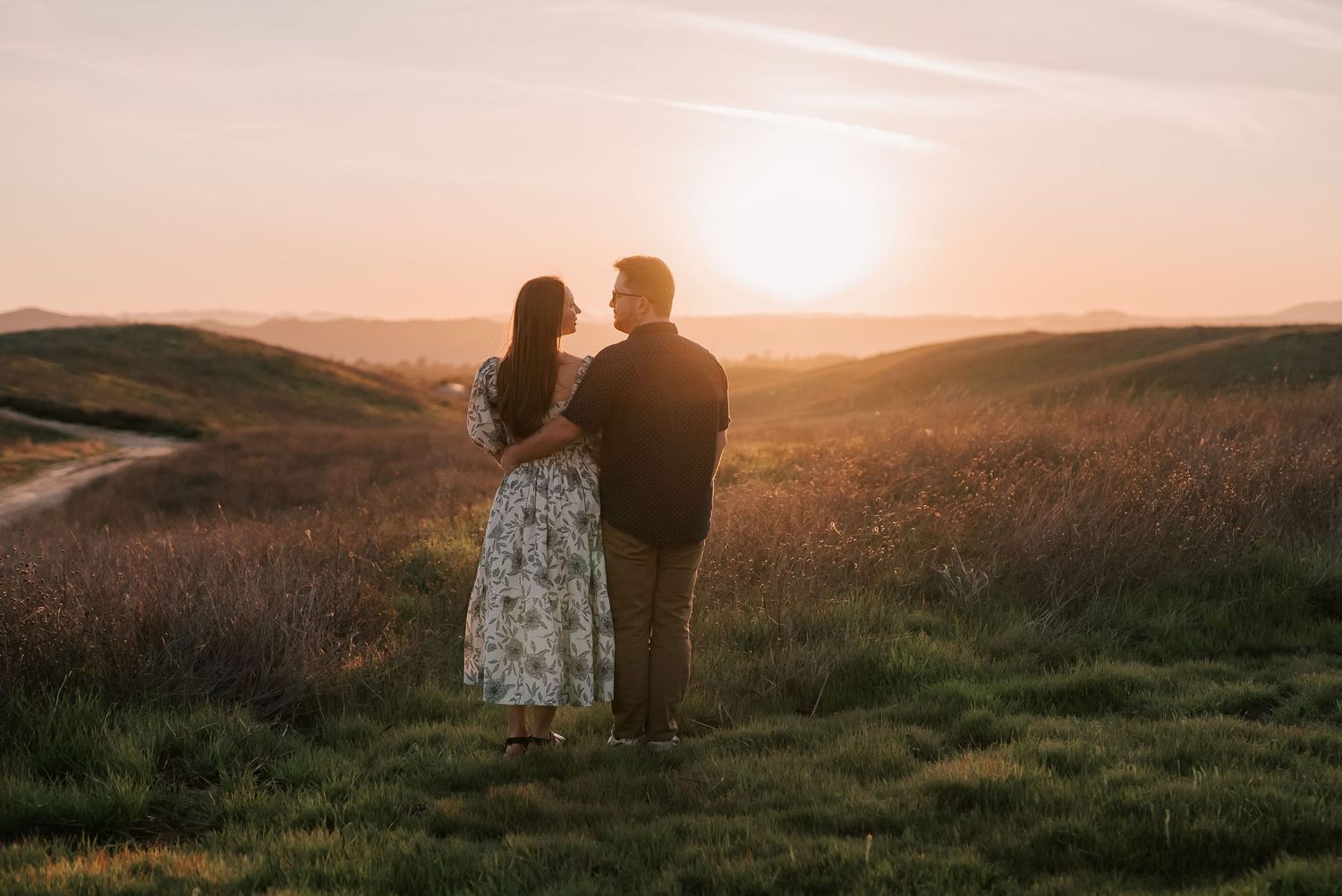 Couple standing together watching a golden sunset over rolling green hills and scenic countryside landscape.