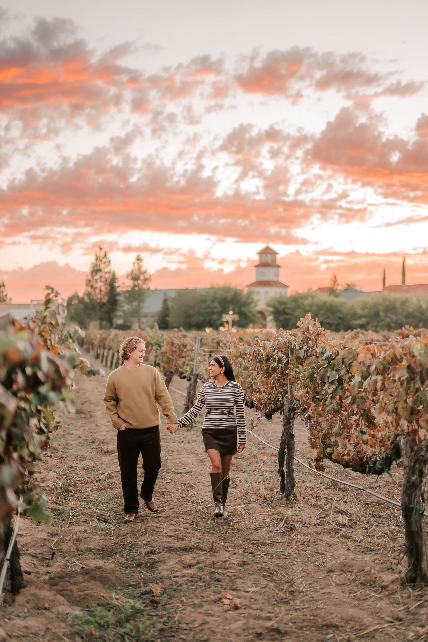 Couple holding hands walking through vineyard rows at sunset with dramatic pink and orange sky.