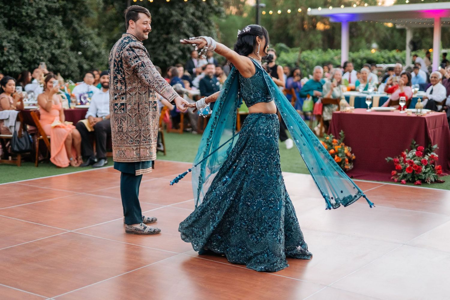 Couple dancing at outdoor Indian wedding, woman in teal embroidered lehenga, man in ornate sherwani.
