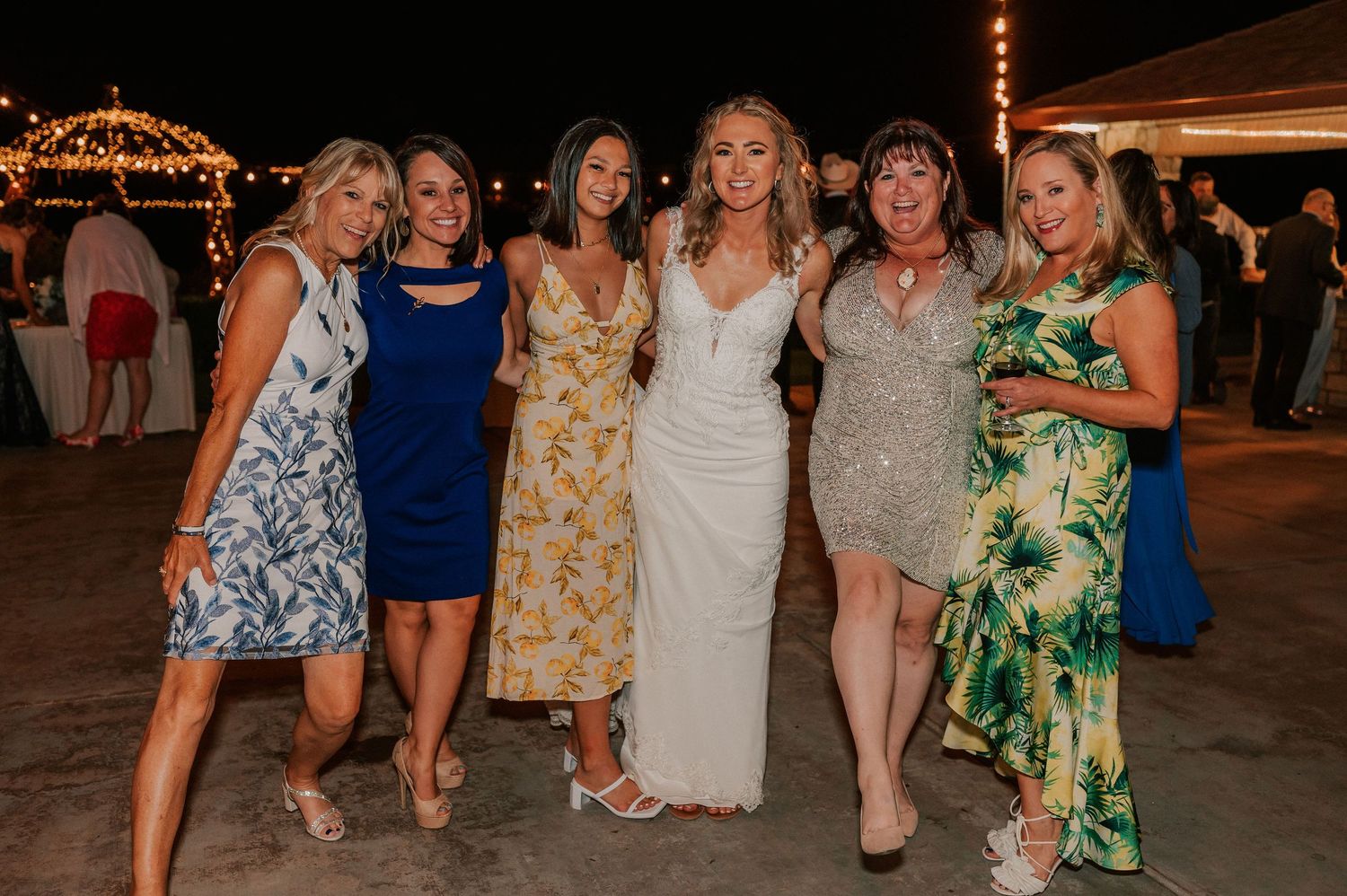 Six women posing together at an outdoor beach wedding celebration at night, one wearing a white bridal gown.