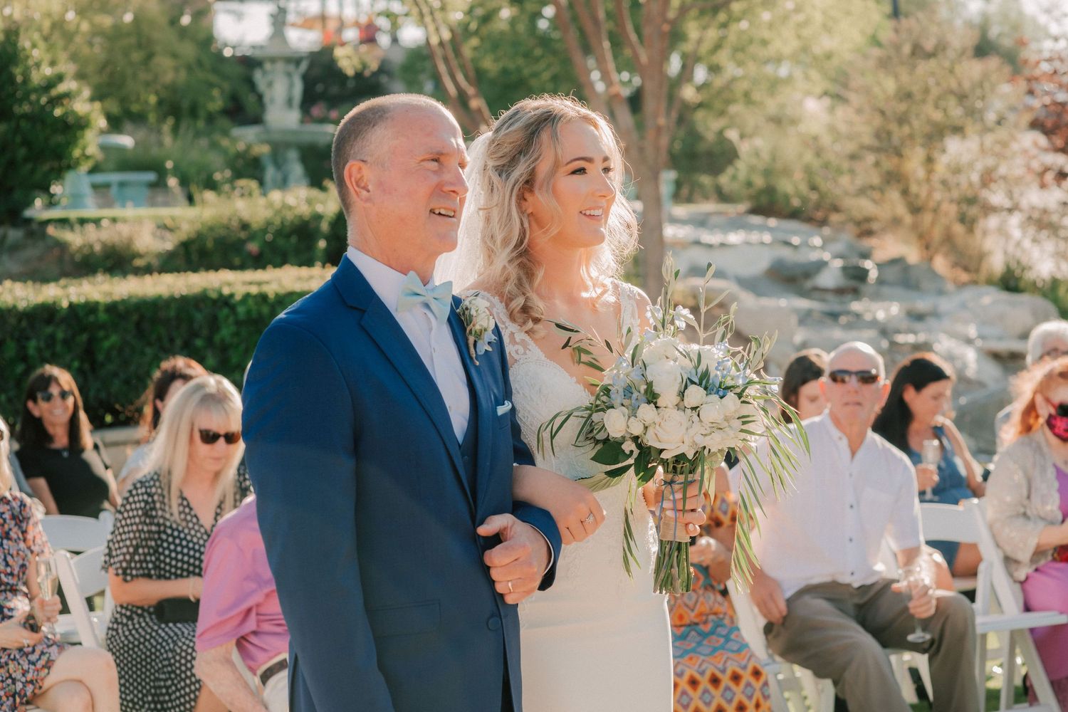 Bride walks down the aisle with her father at an outdoor wedding ceremony, holding a white and green bouquet.