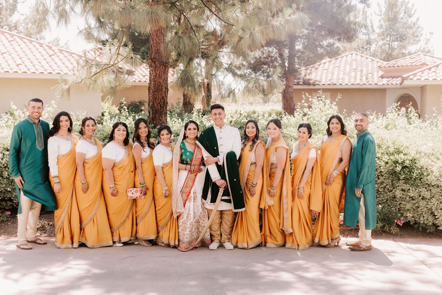 Wedding party poses in matching mustard yellow sarees and teal outfits with bride and groom.