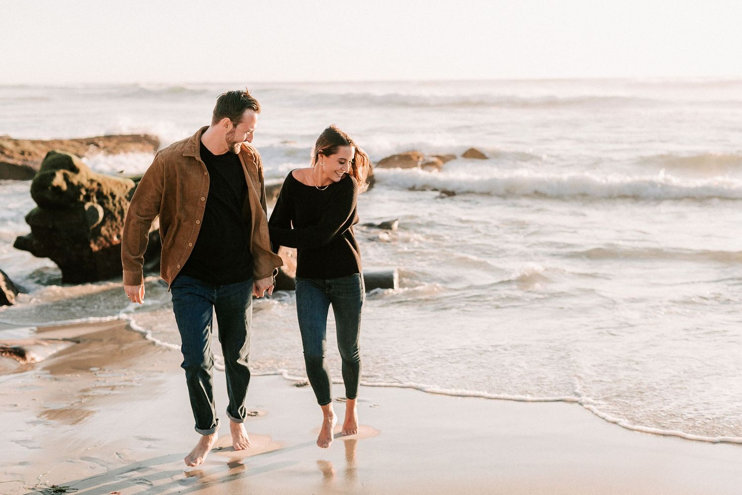 Barefoot couple walking hand in hand along a sandy beach with gentle waves at golden hour.