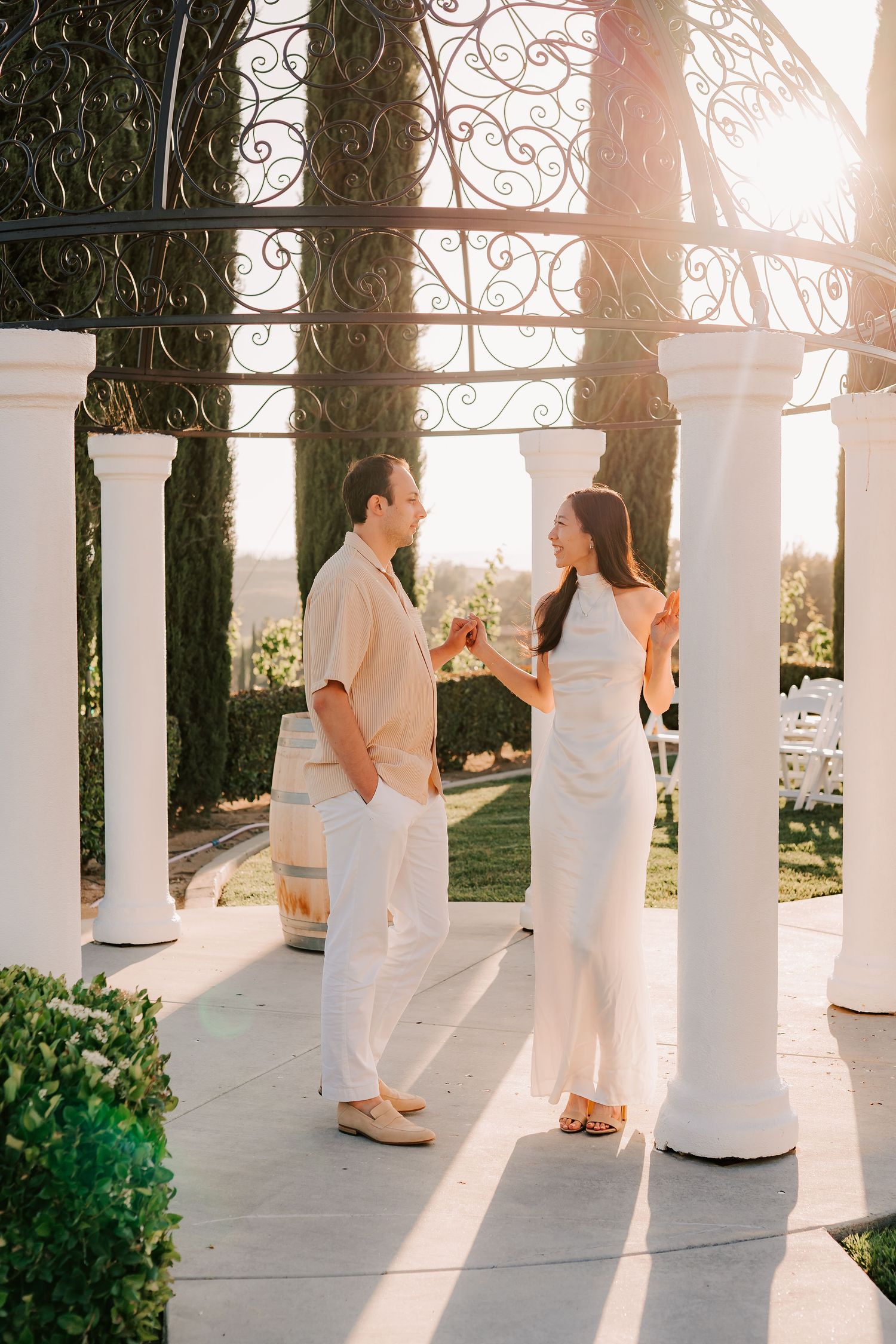 Couple in white attire toasting with wine glasses under elegant iron pergola with white columns at golden hour.