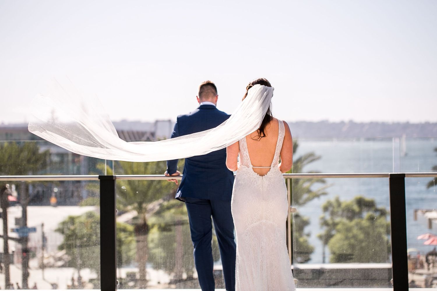 Bride and groom stand on a rooftop balcony overlooking the ocean, her veil flowing in the breeze.