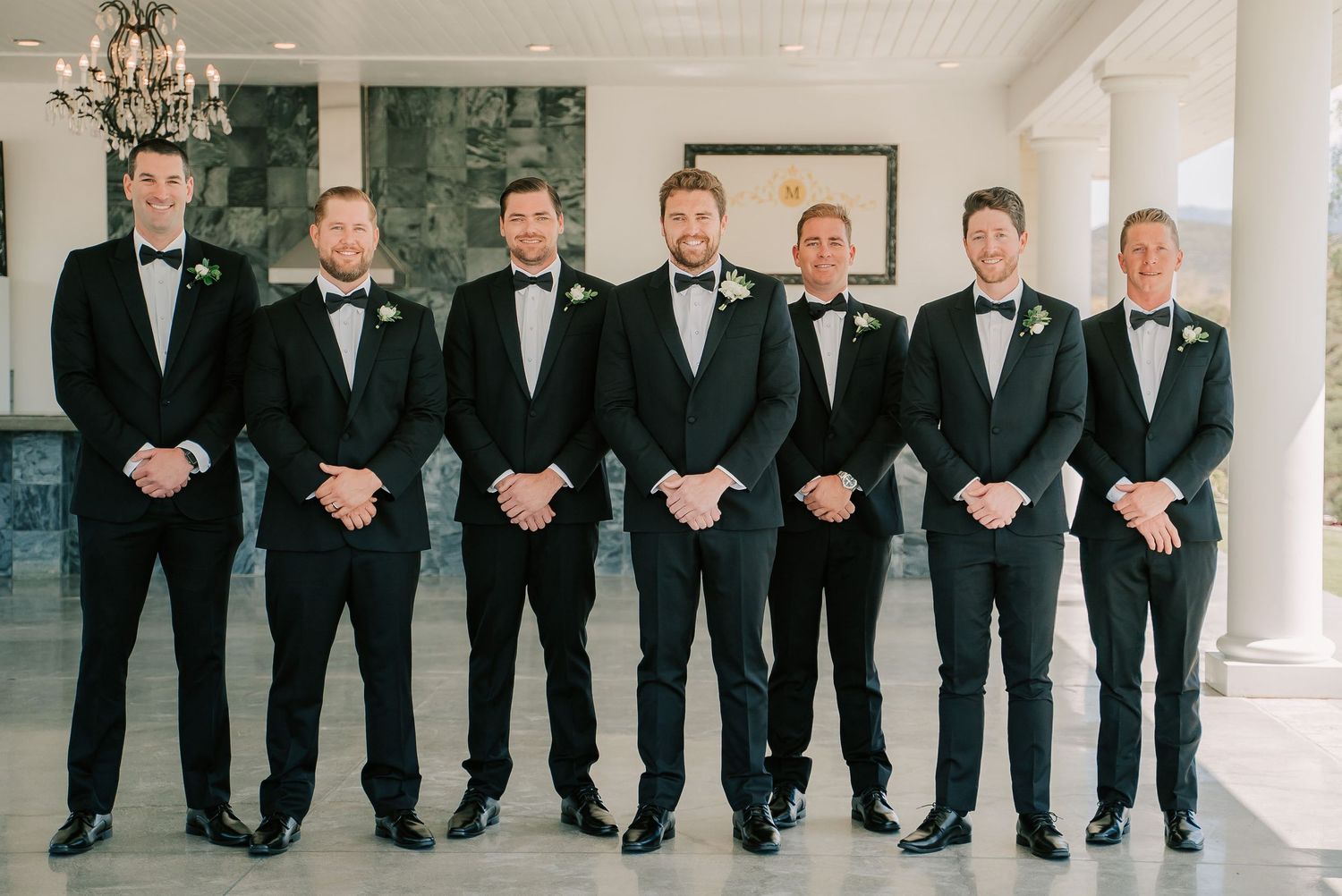 Seven groomsmen in black tuxedos and bow ties with white boutonnieres pose in a line at an elegant wedding venue.