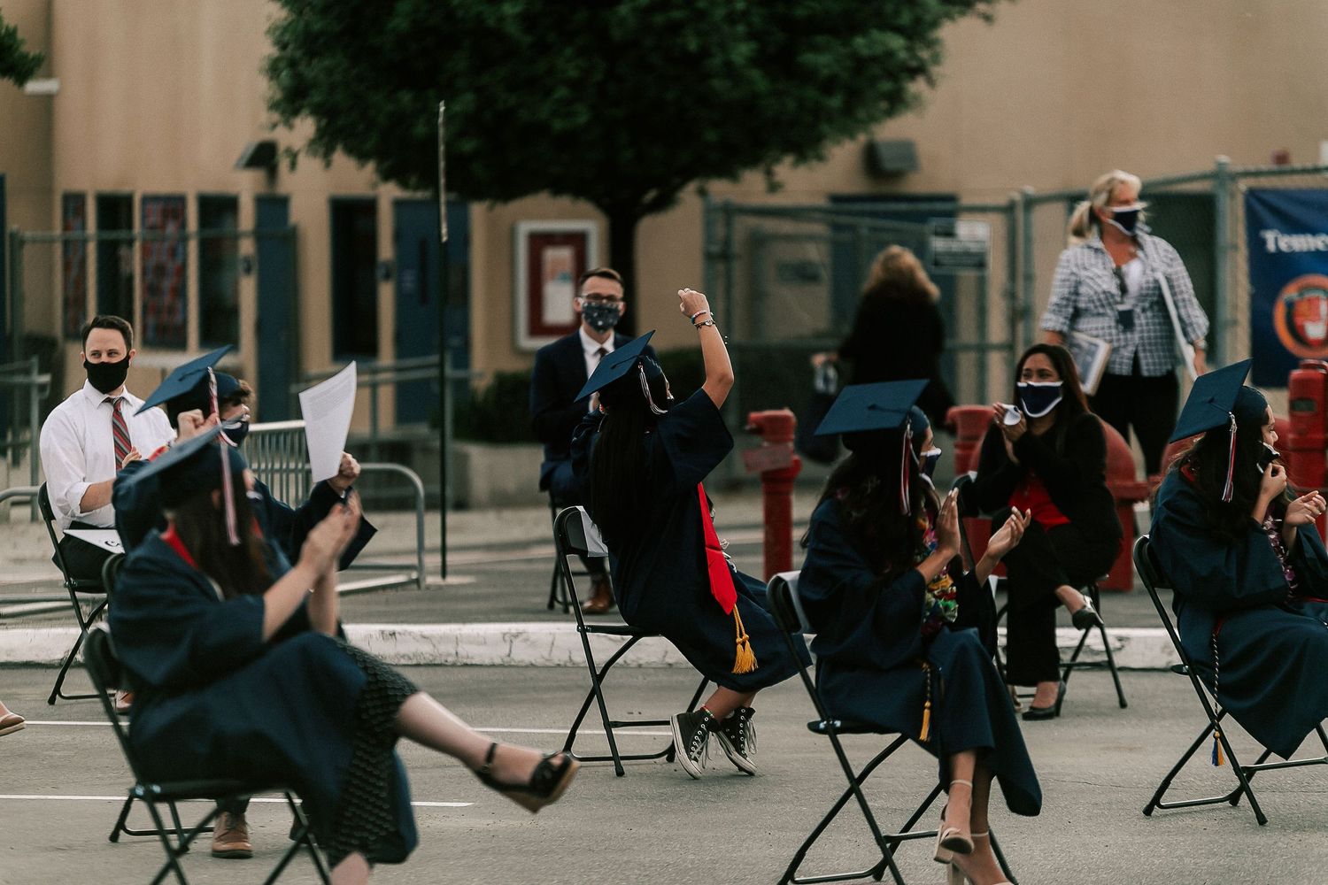 Graduates in caps and gowns celebrate at an outdoor socially distanced graduation ceremony, wearing face masks.