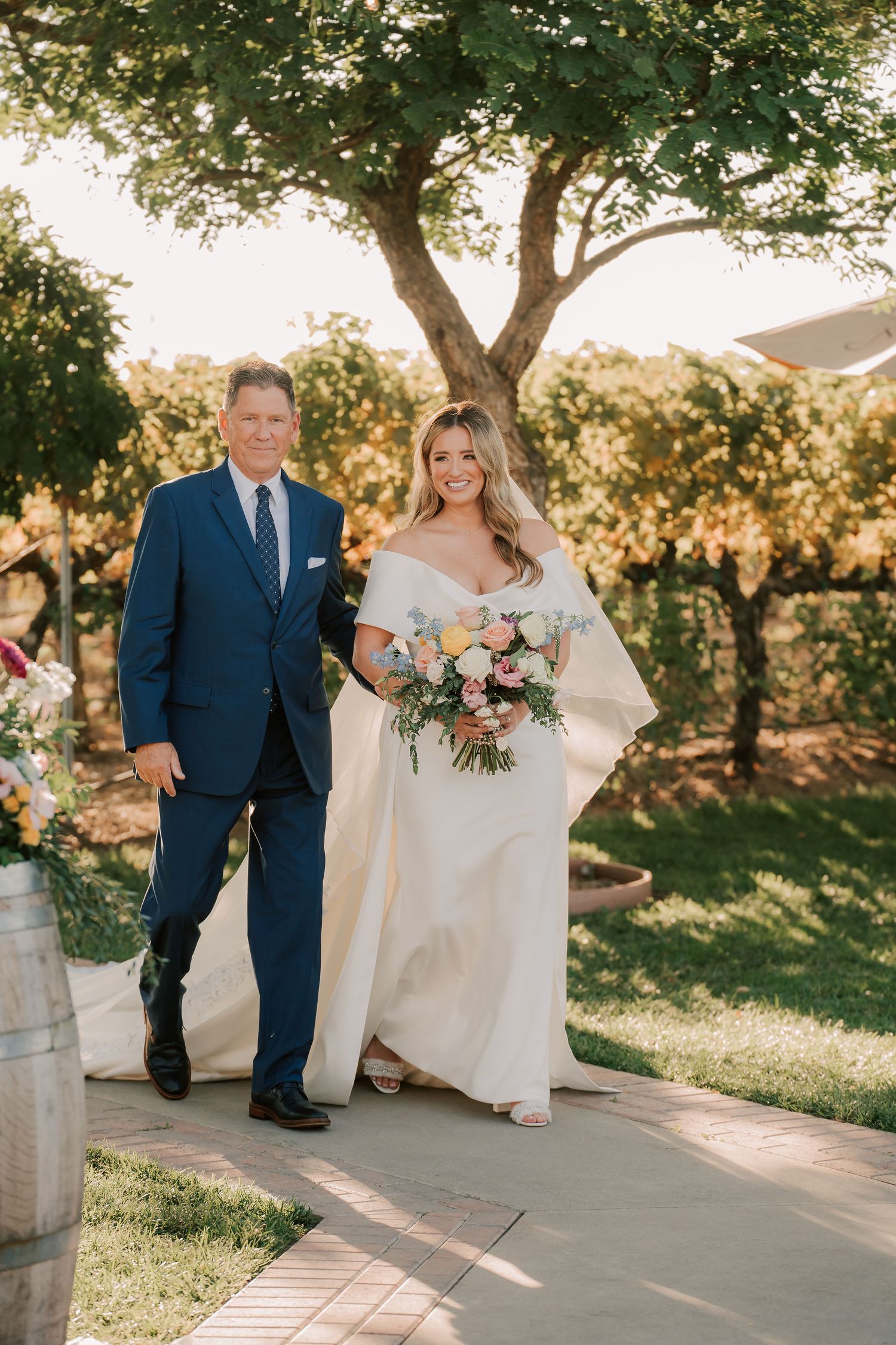 Bride in off-shoulder white gown holding floral bouquet walks with suited man through vineyard wedding aisle.