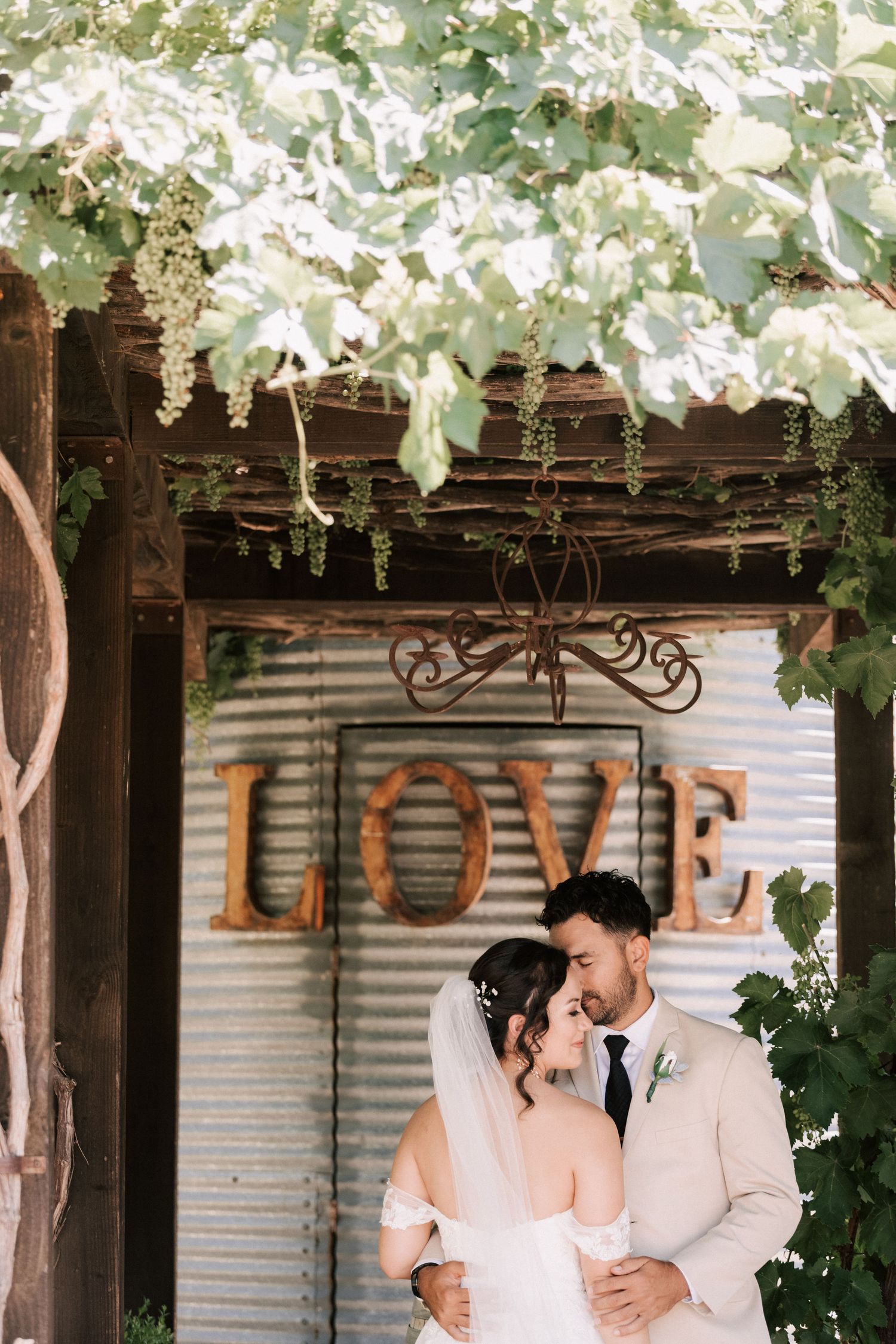 Bride and groom embrace under a rustic LOVE sign surrounded by lush green vines at an outdoor wedding venue.