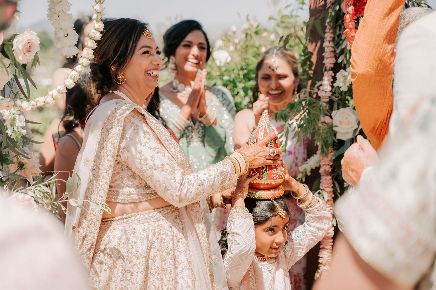 Indian bride in ivory lehenga smiles during traditional Hindu wedding ceremony under floral mandap with guests.