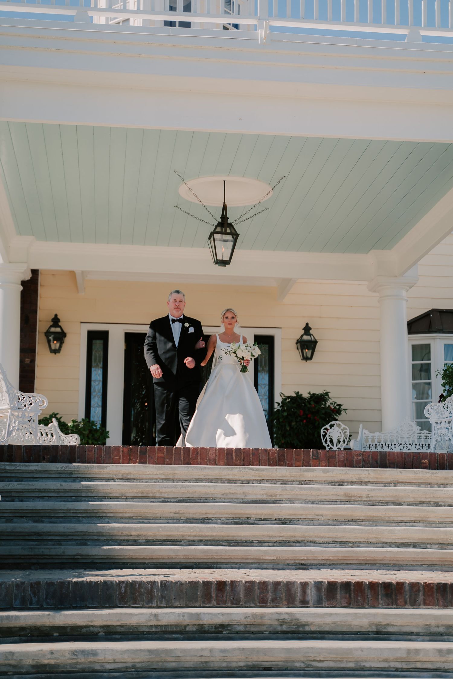 Bride in white gown and groom in tuxedo descend grand brick steps of elegant Southern wedding venue.