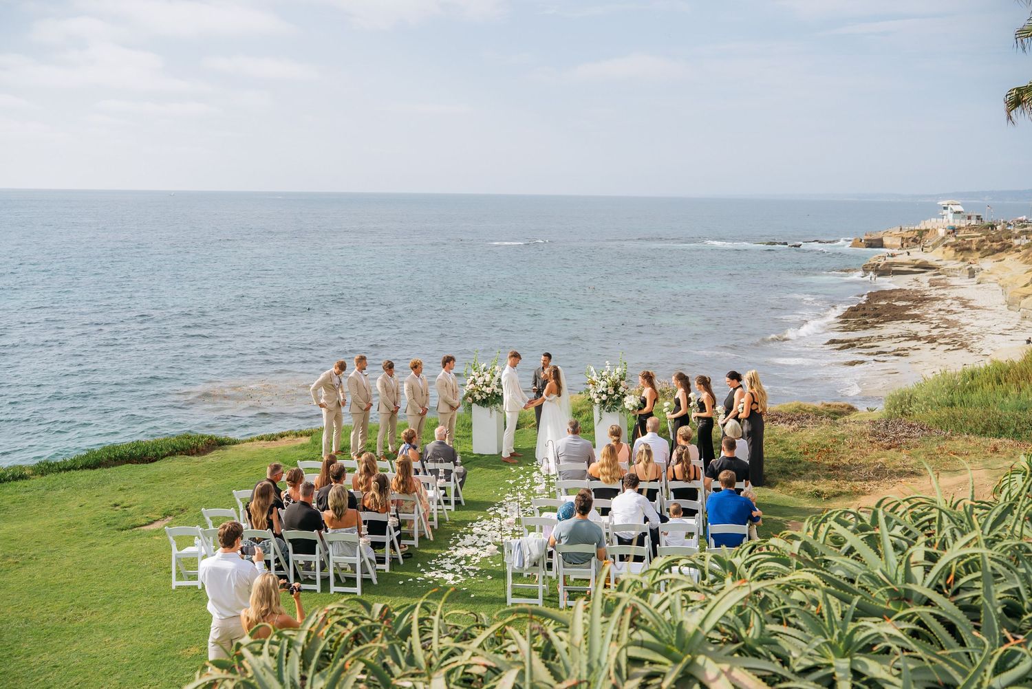 Outdoor beachside wedding ceremony on green cliffs overlooking the ocean with guests seated in white chairs.