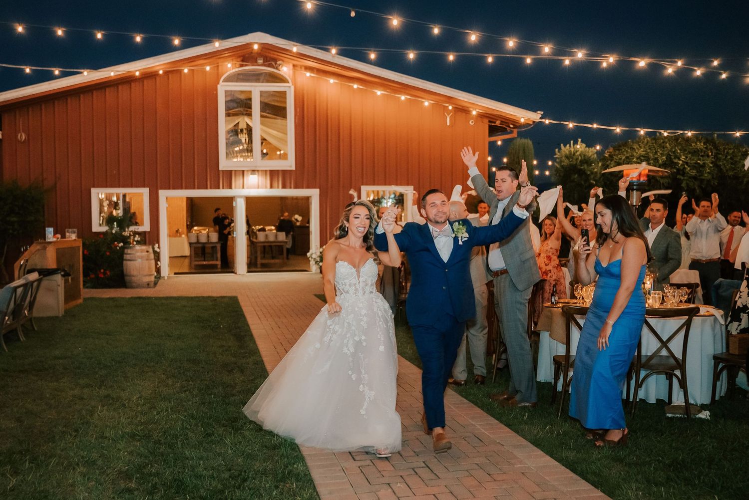 Bride and groom celebrate grand entrance at outdoor barn wedding reception under string lights.