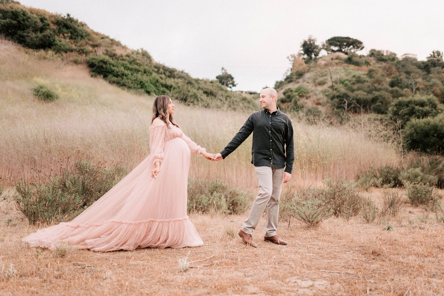 Pregnant woman in pink gown holds hands with man in black shirt in golden field.