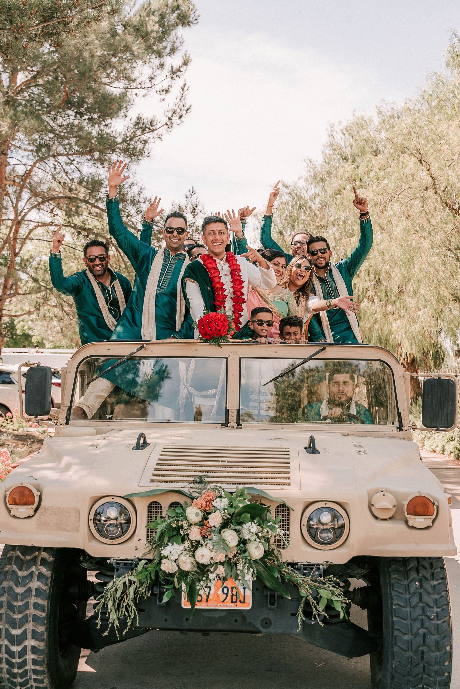 Groom and groomsmen in teal sherwanis celebrating atop a decorated military Humvee.