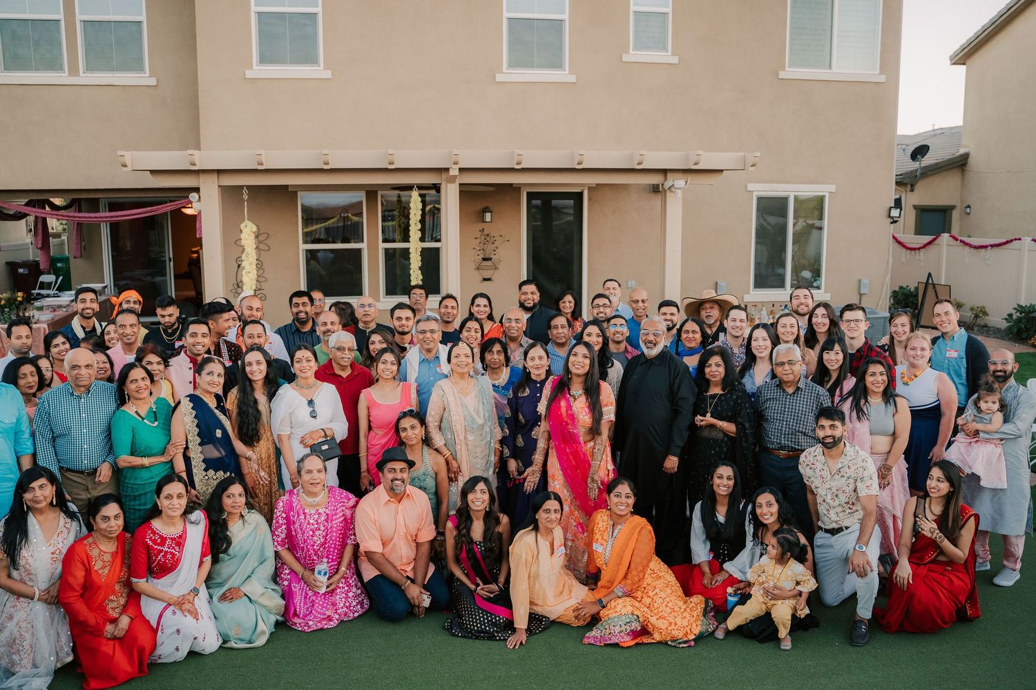 Large Indian wedding family group photo outdoors in front of a beige house with guests in colorful traditional attire.