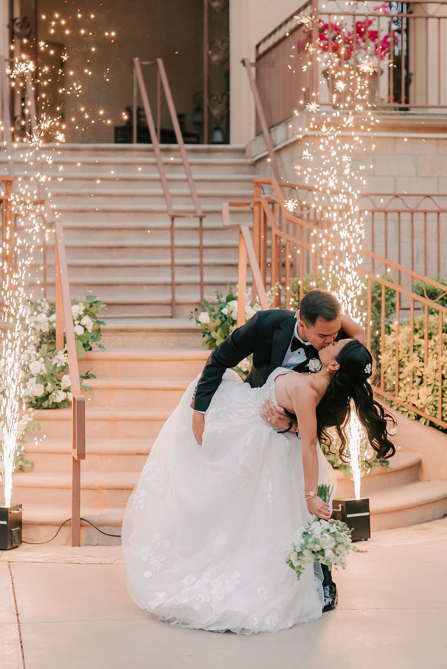 Groom dips bride for a romantic kiss in front of sparkling fountain fireworks on elegant staircase at wedding reception.