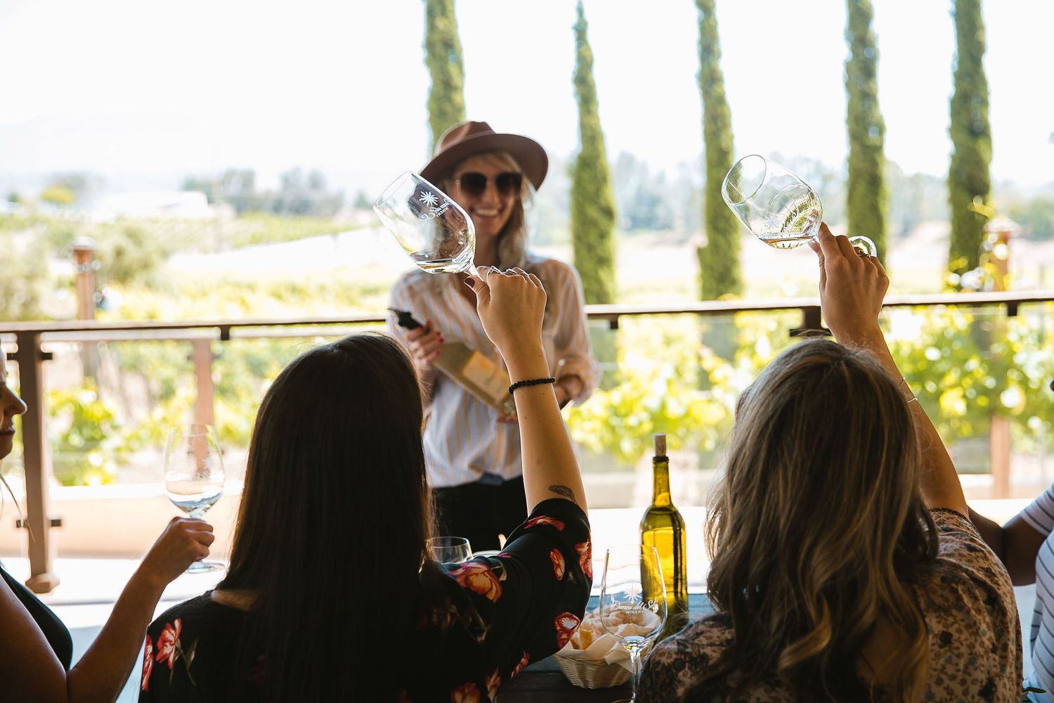 Women toasting wine glasses at a vineyard winery tasting, with lush green vines and a smiling server in a hat.