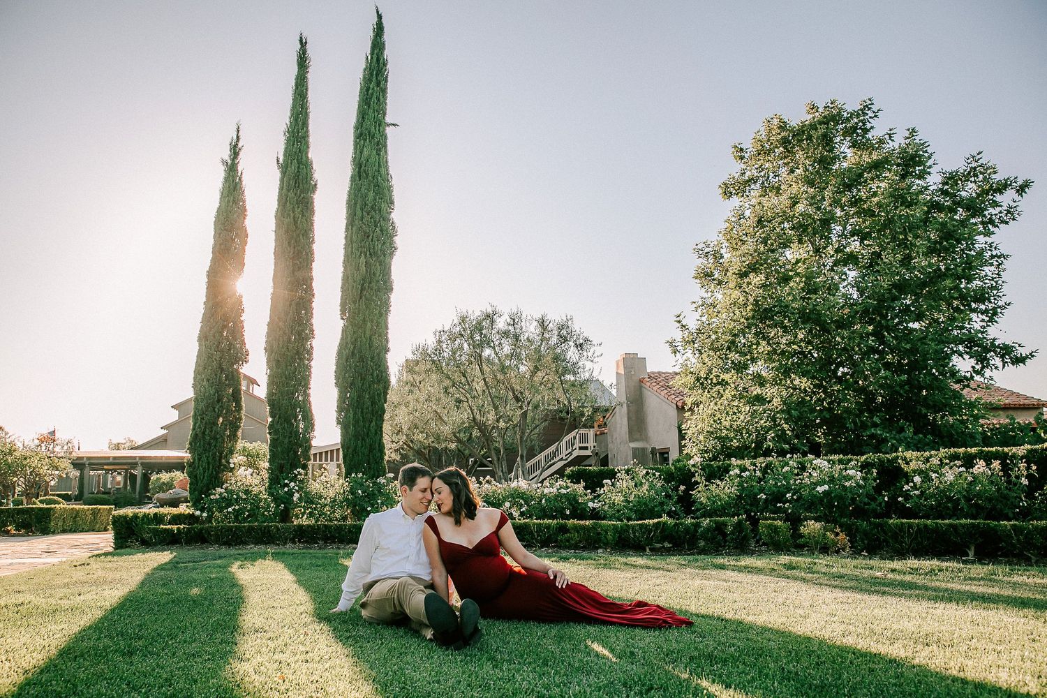 Couple in elegant attire relaxing on lush green lawn with tall cypress trees and sunlit estate in background.