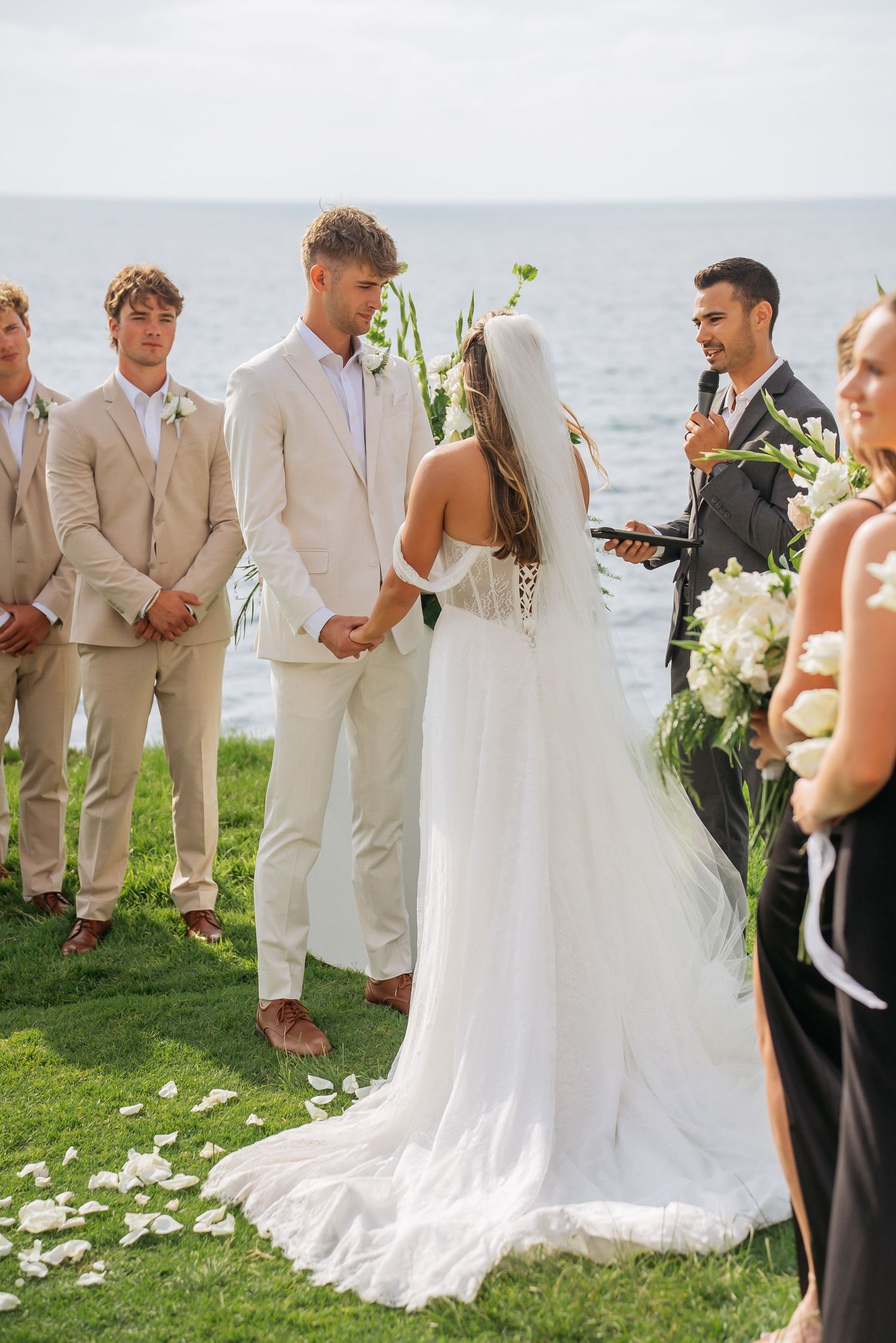 Bride and groom exchange vows at oceanfront ceremony with groomsmen and floral arrangements nearby.