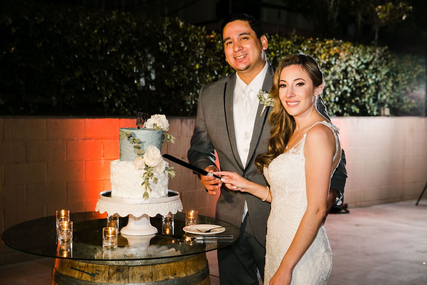 Bride and groom smiling while cutting their elegant white wedding cake outdoors at a romantic evening reception.