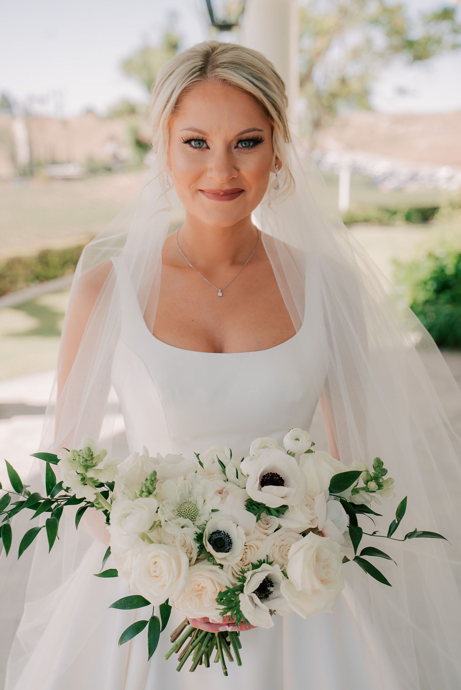 Bride in white square-neck gown holding white anemone bouquet with greenery, wearing veil and delicate necklace.