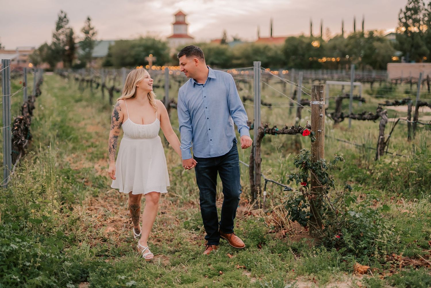 Couple holding hands walking through vineyard rows at sunset with historic building in background.