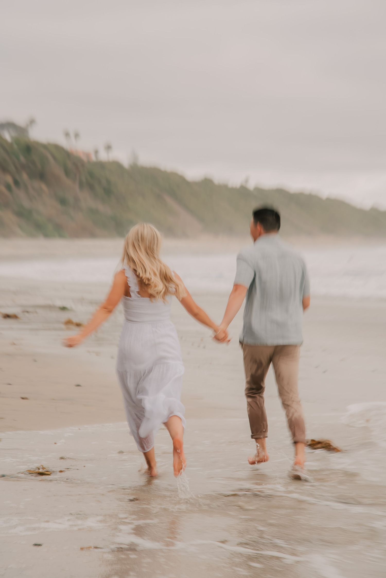 A couple holds hands running barefoot along a misty beach, woman in white dress and man in sage shirt.