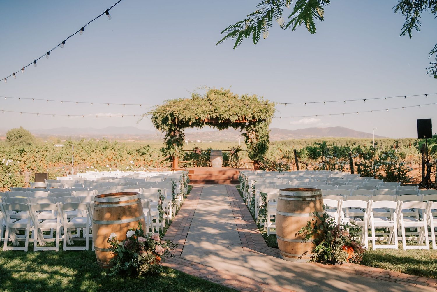 Outdoor vineyard wedding ceremony setup with white chairs, barrel decor, floral arch, and string lights under a clear blue sky.