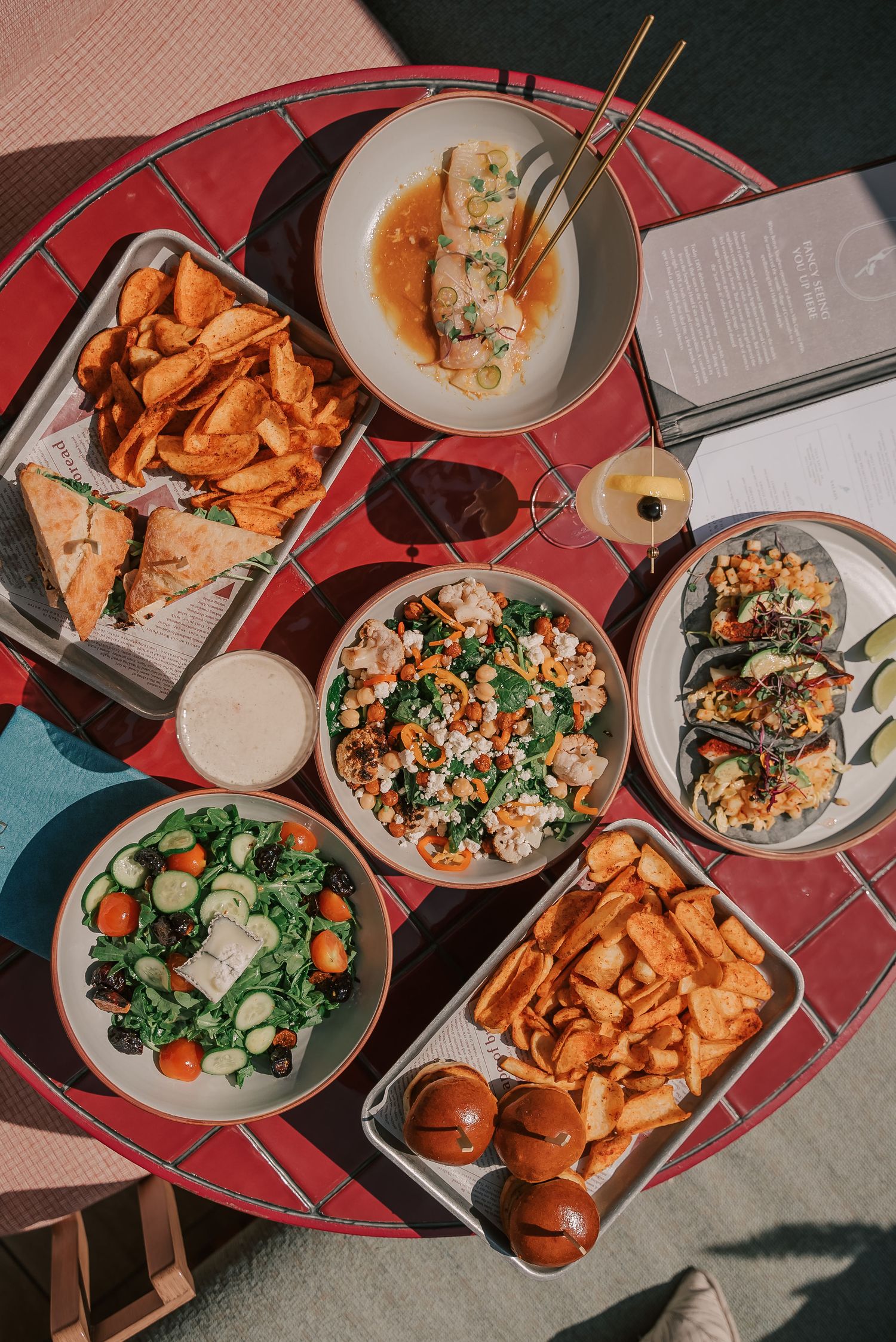 Overhead view of a vibrant spread of dishes on a red table, including salad, chips, grain bowl, tacos, and sliders.