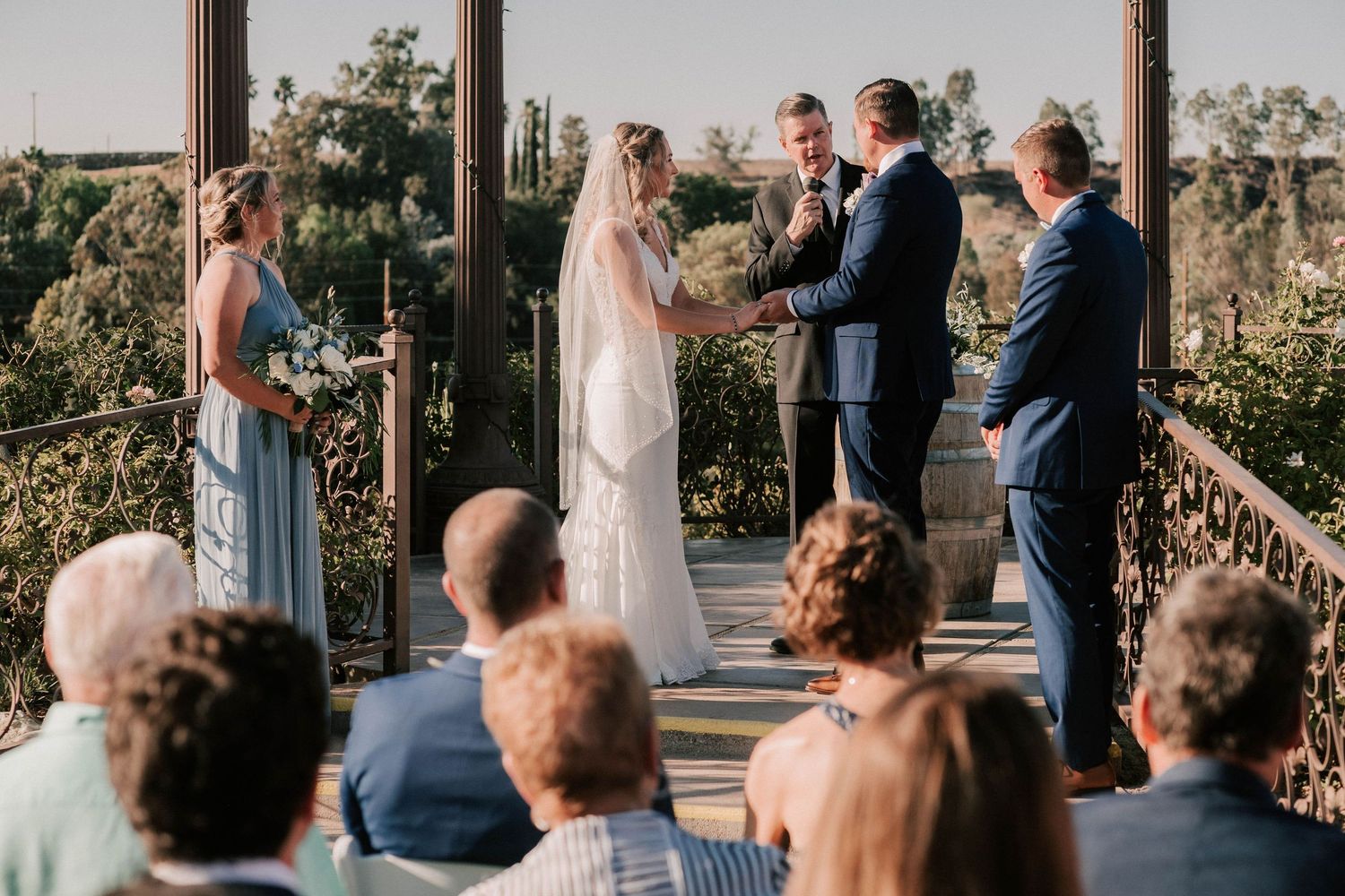 Outdoor wedding ceremony on a wooden terrace with bride, groom, officiant, and bridesmaid before seated guests.