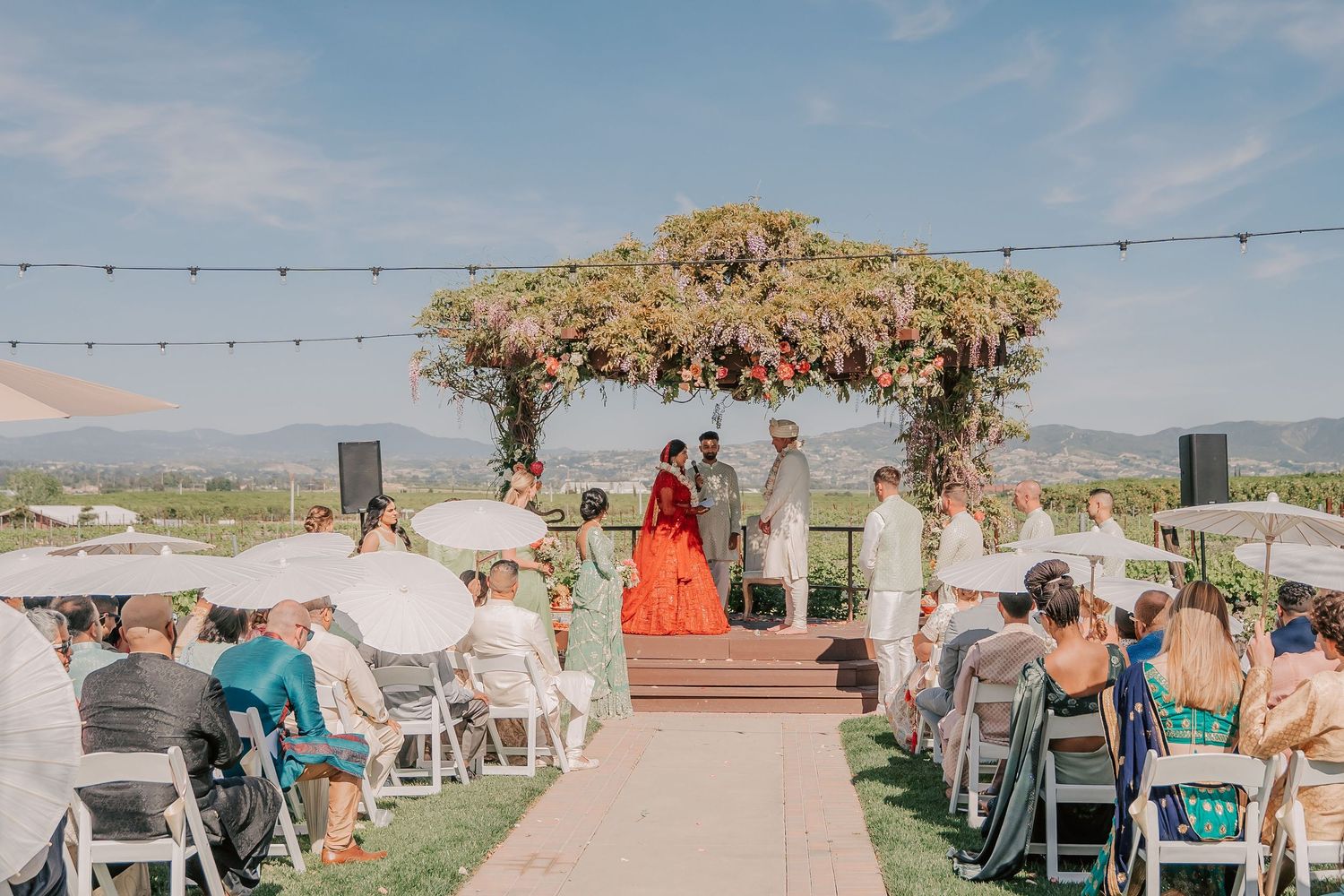 Colorful Indian wedding ceremony under floral mandap with bride in red lehenga at outdoor vineyard venue.