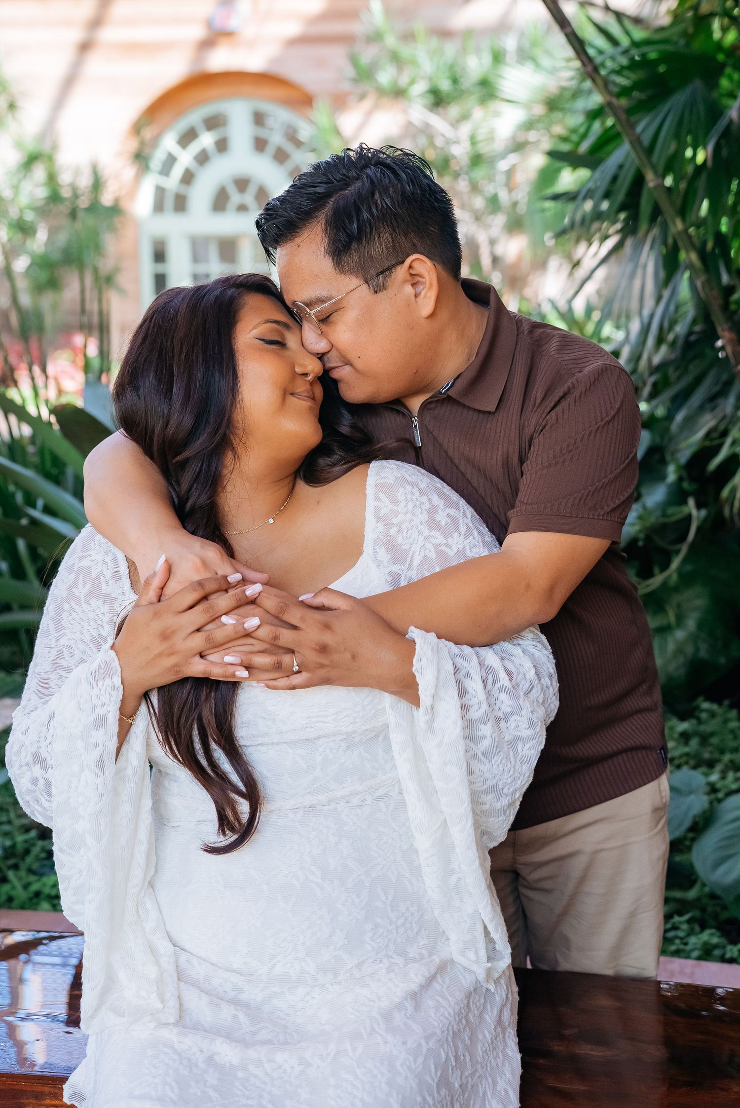 A couple shares a tender embrace outdoors, woman in white lace dress, man in brown polo, surrounded by lush greenery.