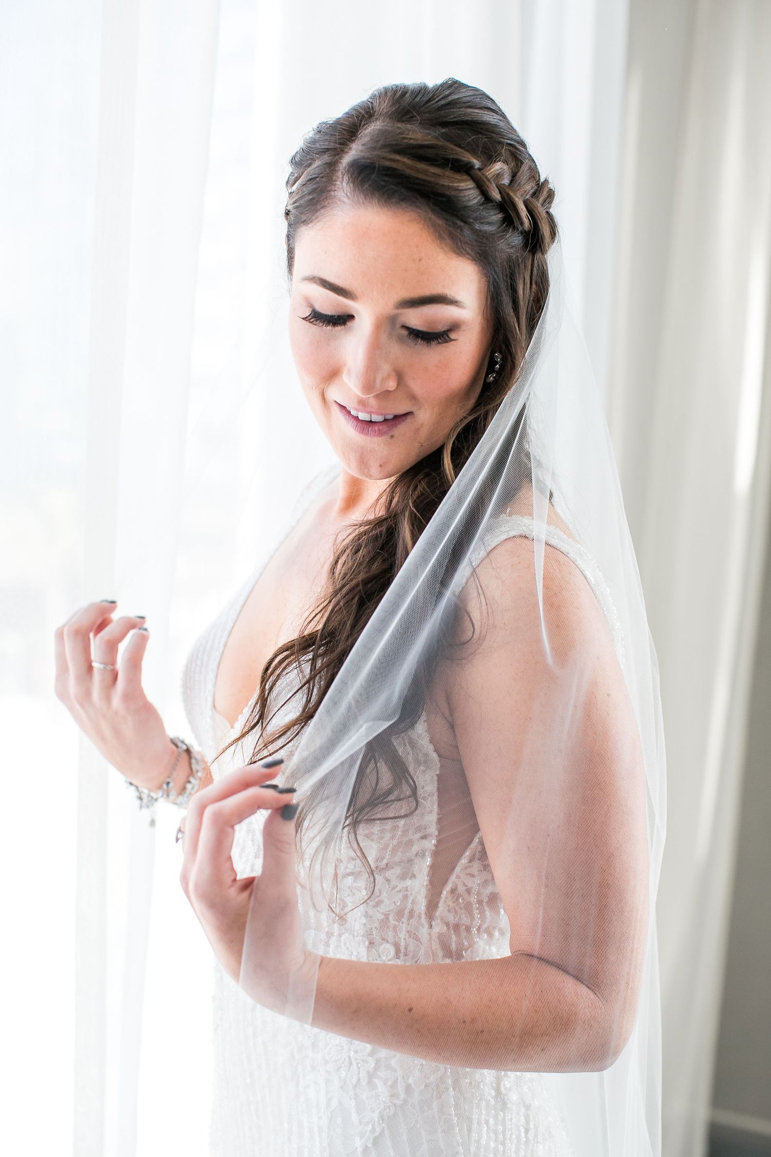 Bride in lace wedding dress and veil smiling softly near a bright window on her wedding day.