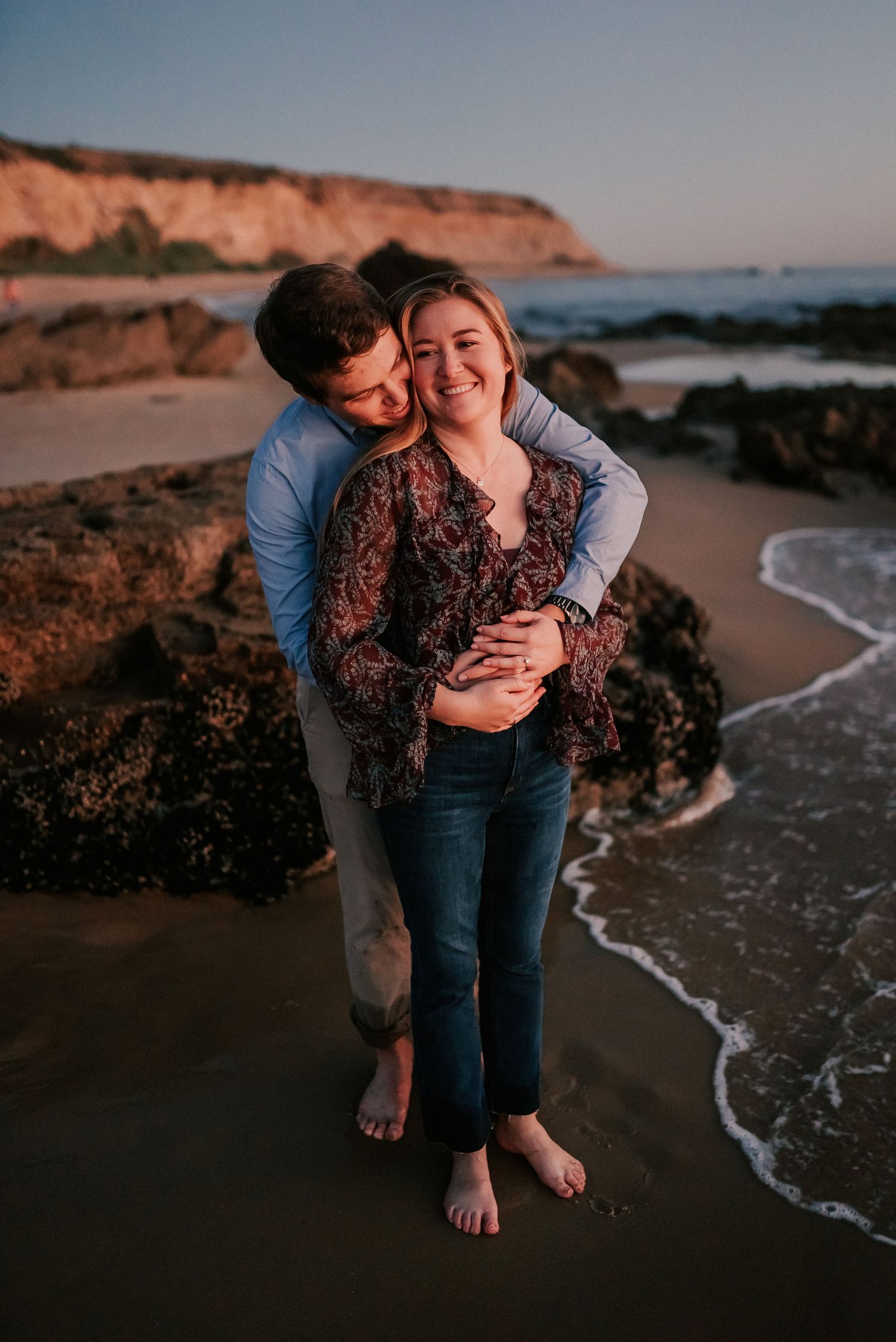 Couple embracing on a rocky beach at sunset, woman smiling as man hugs her from behind near ocean waves.
