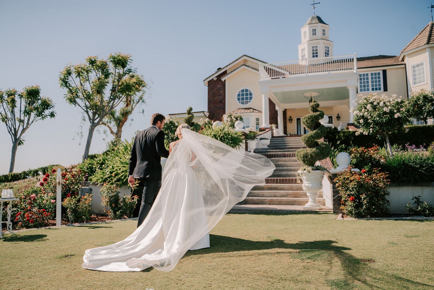 Bride and groom walking on green lawn outside elegant wedding chapel with flowing veil billowing in the breeze.