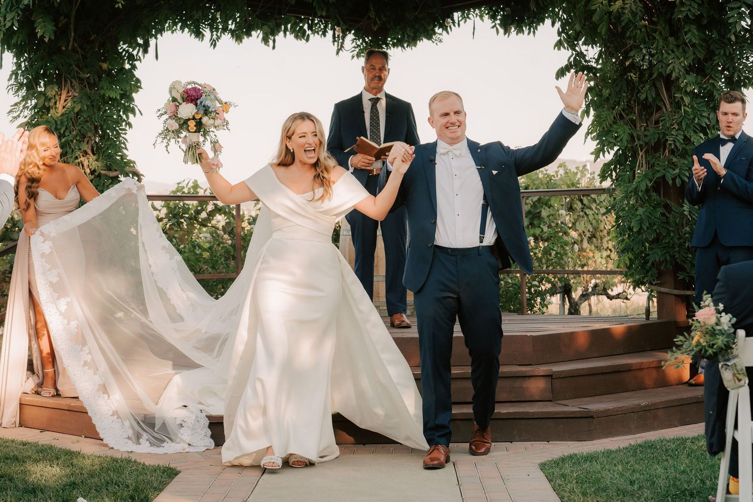 Joyful bride and groom exit wedding ceremony holding hands, bride in elegant white gown, groom in navy suit.