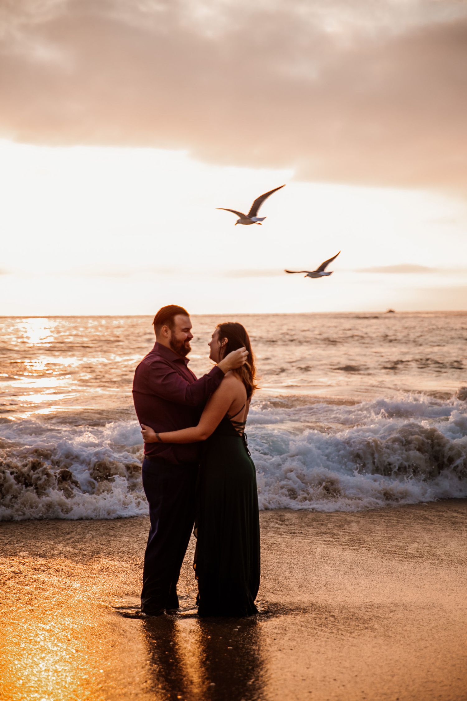 Couple embracing at sunset on a beach with waves and seagulls flying overhead in golden light.