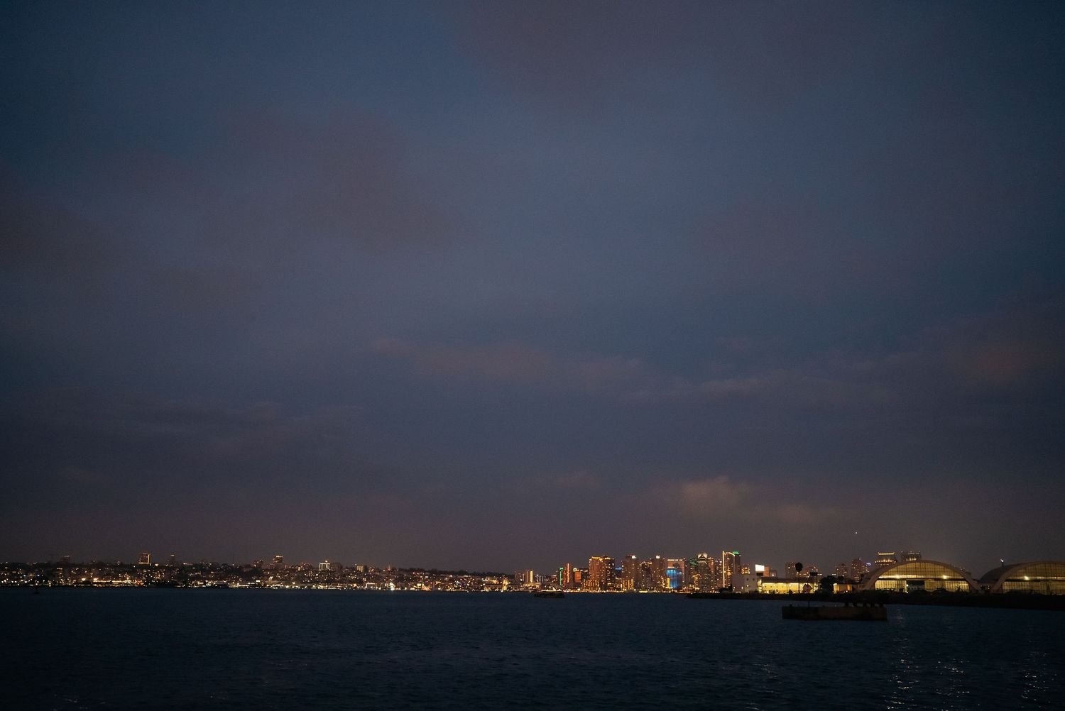 Stunning nighttime cityscape skyline reflecting over calm dark ocean waters under cloudy sky.