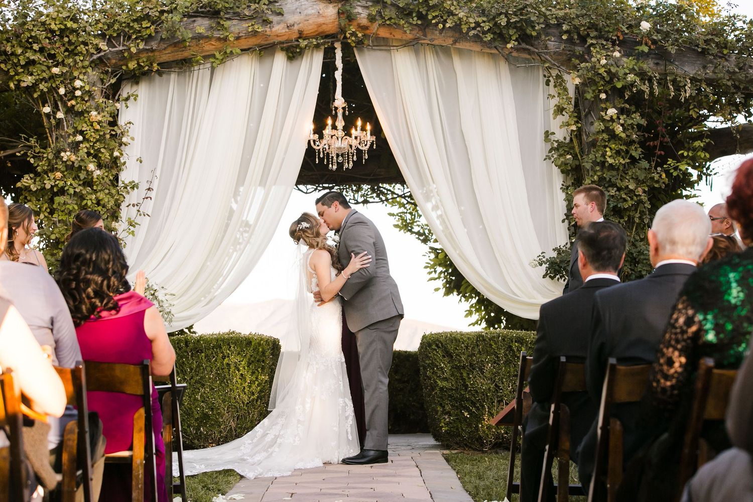 Bride and groom share first kiss under floral chandelier arch with white draping at outdoor garden wedding ceremony.