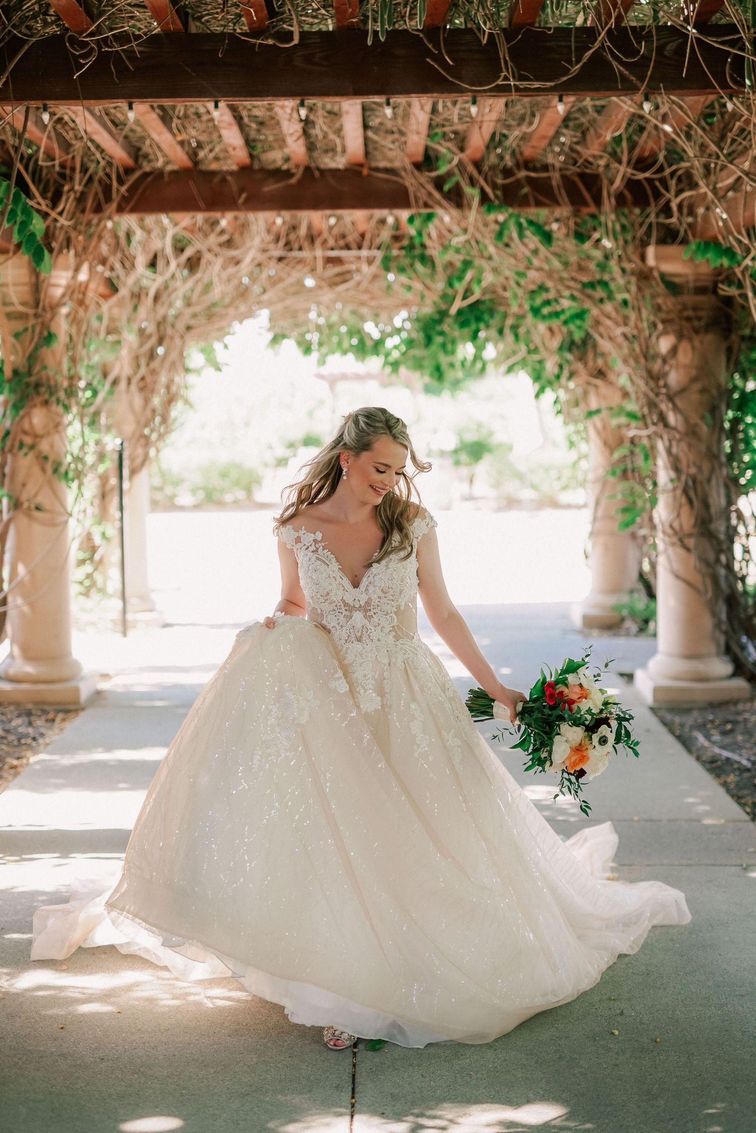 Bride in lace ballgown twirling under vine-covered pergola, holding colorful bouquet in sunlit outdoor setting.